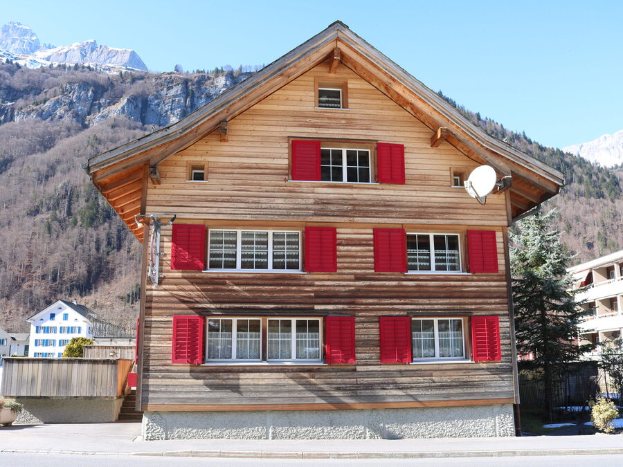 Giebelseite des Hauses mit roten Fensterläden und Bergkulisse